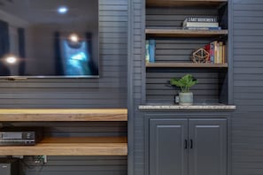 Un salon moderne avec un téléviseur à écran plat fixé au mur, des étagères en bois ornées de livres et d’objets décoratifs, et une petite plante en pot posée sur un comptoir.