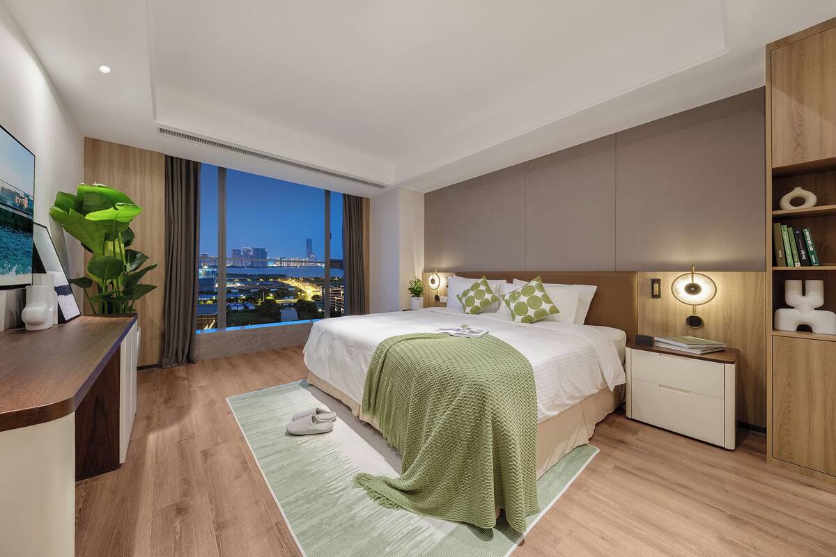 Une chambre d’hôtel moderne dotée d’un grand lit, de tables de chevet, d’un bureau et offrant une vue sur le paysage urbain.