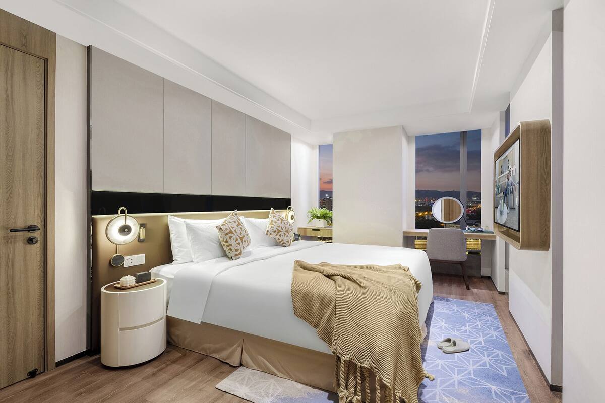 Une chambre d’hôtel moderne avec un grand lit, une table de chevet, une lampe et une vue sur la ville au crépuscule.