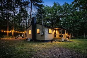 Une cabane avec une véranda, une cheminée et des guirlandes lumineuses, située dans un cadre forestier.