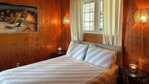 Una habitación con paneles de madera, una cama, un cuadro de una escena de playa y dos lámparas fijadas a la pared.