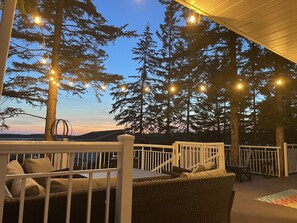 Une terrasse extérieure aménagée avec des meubles en osier et des guirlandes lumineuses, entourée de grands arbres et surplombant un ciel dégagé.