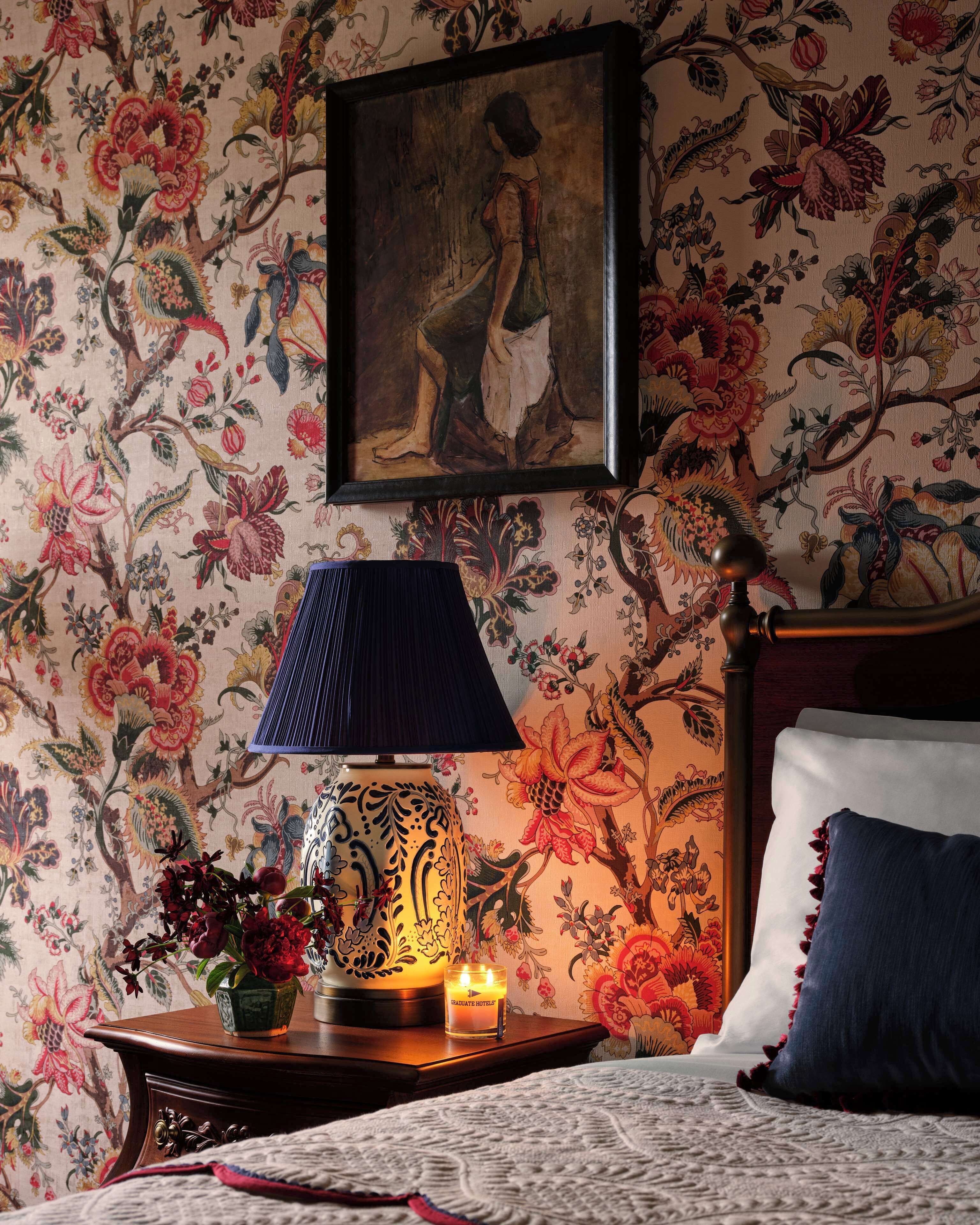 A bedroom with floral wallpaper, a bed with a blue pillow, a nightstand with a lamp and a candle, and a painting above the bed.