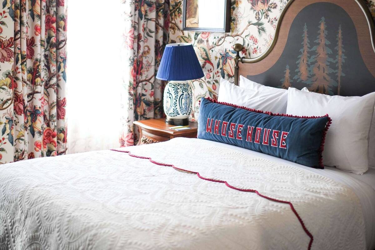 Un dormitorio con papel tapiz floral, una cama con cubrecama blanco, una lámpara azul y un almohadón con el texto "WHOSE HOUSE" bordado.