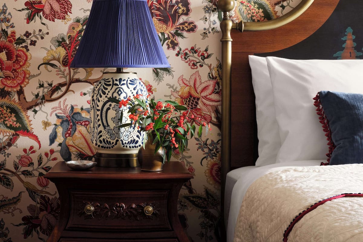 Un dormitorio con papel tapiz floral, un libro enmarcado en la pared, una mesita de noche con lámpara y una cama con almohadas.