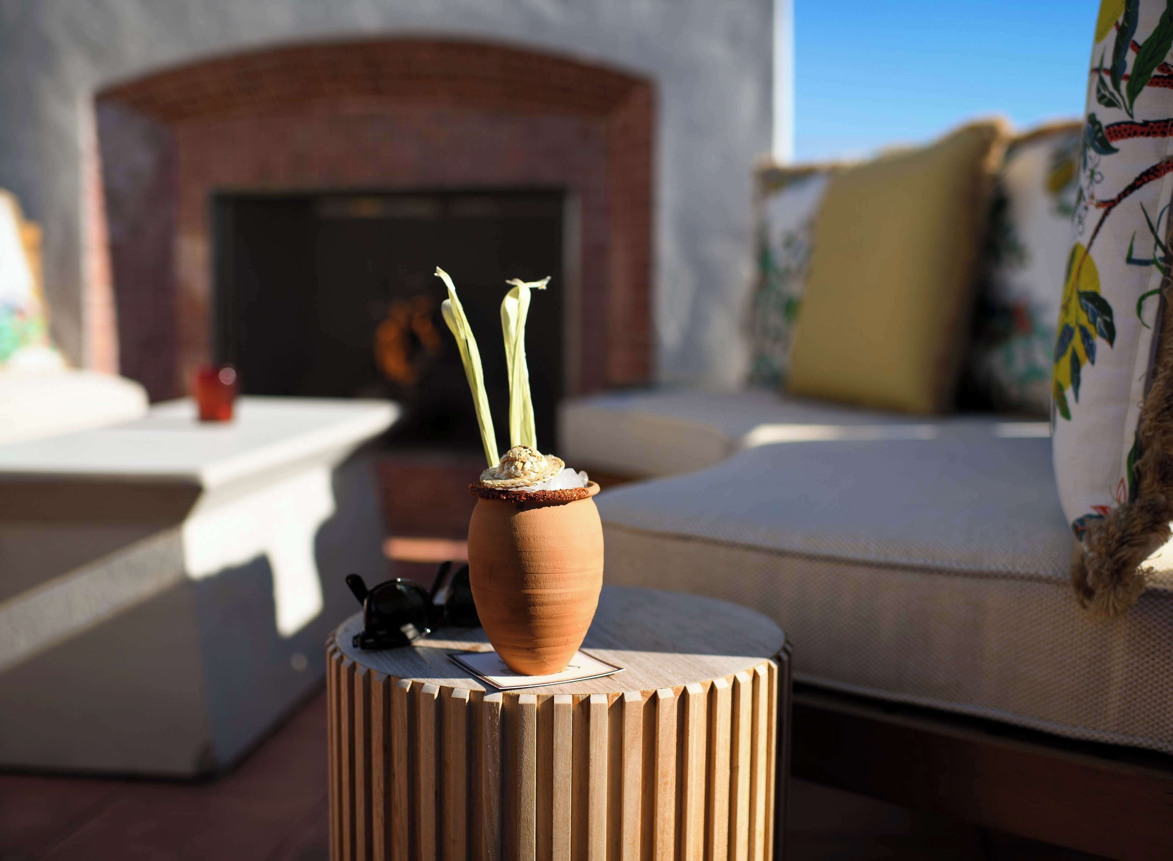 A terracotta pot with a decorative item on a wooden stool in a sunlit outdoor seating area with a brick fireplace in the background.
