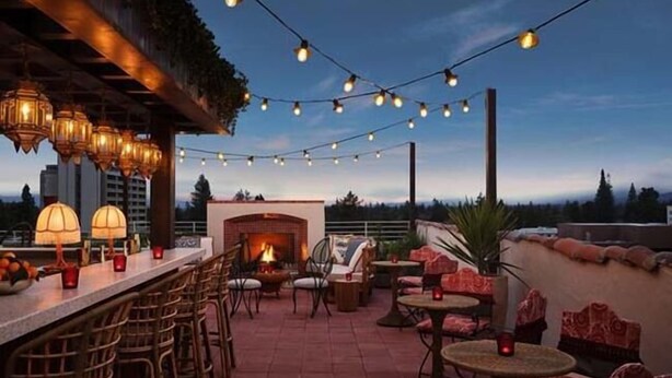 An outdoor patio with a brick fireplace, string lights, wicker furniture, and a view of the city skyline. - Graduate by Hilton Palo Alto (Palo Alto)