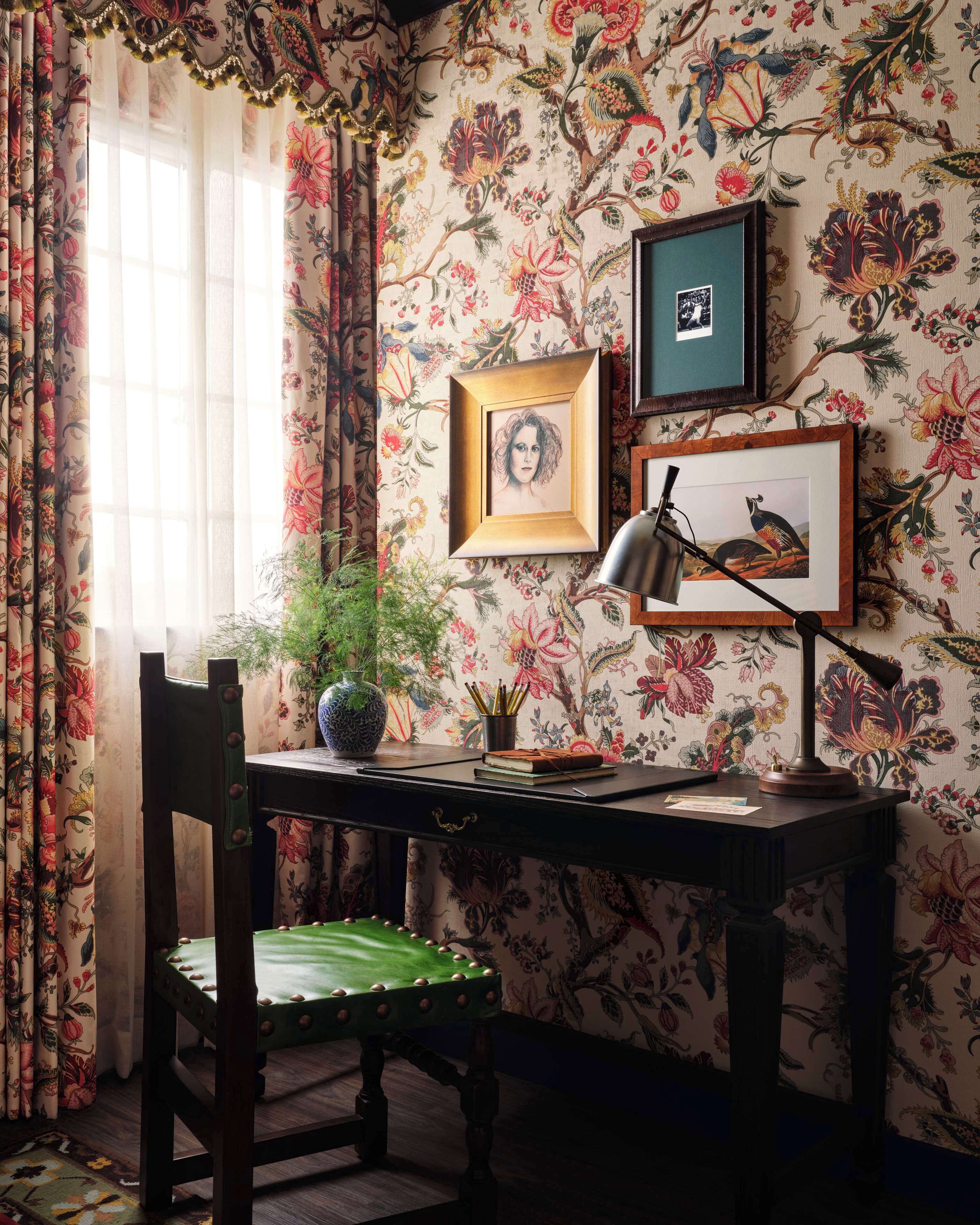 A room with floral wallpaper, a desk with a lamp, framed pictures, and a chair.