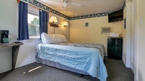 Een slaapkamer met een bed, nachtlampjes, een bureau met een koffiezetapparaat, een raam met gordijnen, een plafondventilator en een kleine koelkast.
