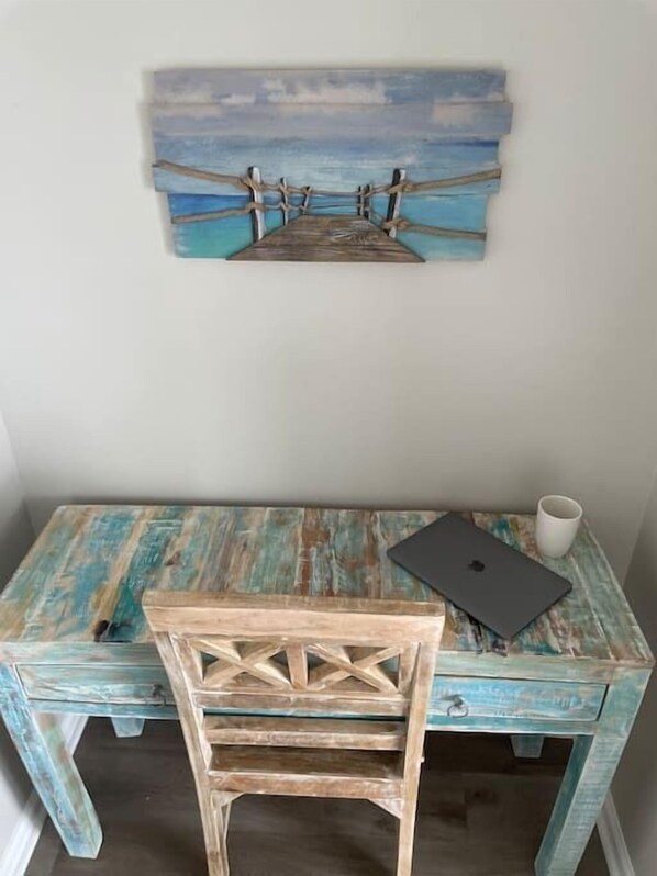 Un bureau en bois rustique, sur lequel se trouvent un ordinateur portable et une tasse, et un tableau représentant une jetée accroché au mur.