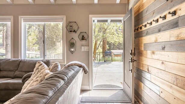 Un salon chaleureux avec un canapé en cuir, des poutres apparentes en bois au plafond et une vue, par une porte, sur un espace extérieur équipé d’un barbecue.