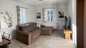 A modern living room with a brown sectional sofa, a wooden coffee table, and a flat-screen TV on a stand. There are framed pictures on the walls, a window with a view of trees, and a radiator under the window.