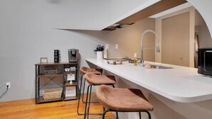 Une cuisine compacte dotée d’un plan de travail blanc, d’un évier encastré et d’une machine à café. On y trouve trois tabourets et une petite étagère contenant des ustensiles de cuisine.