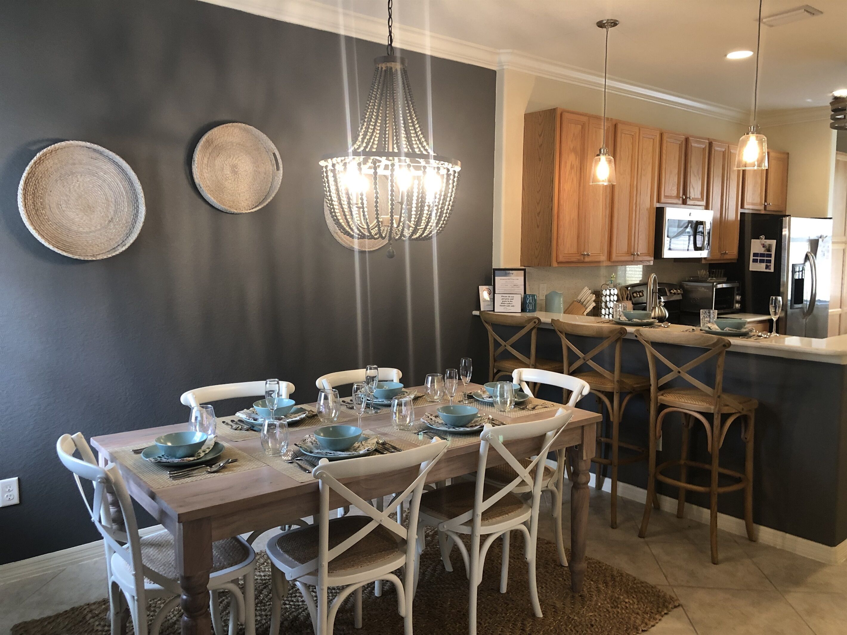 A dining area with a wooden table set for six, surrounded by white chairs. A chandelier hangs above, and wooden cabinets are visible in the background.