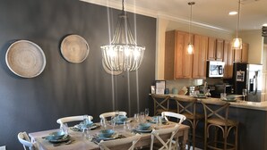 Comedor con mesa de madera dispuesta para seis personas, rodeada de sillas blancas. Un candelabro cuelga encima y se ven armarios de madera al fondo.