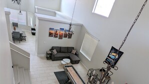 Amplia sala de estar con sofá modular, una larga mesa de comedor y una mesa de billar. Varias ventanas proporcionan luz natural, y se observa un ventilador de techo.