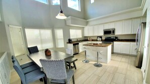 Una cocina moderna con armarios blancos, una isla central con taburetes, una mesa de comedor con sillas y una lámpara colgante.