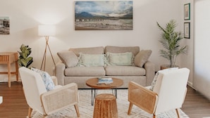 Sala de estar con un sofá color beige, dos sillones blancos, una mesa de centro y una lámpara de pie. Hay un cuadro en la pared y una planta en maceta.