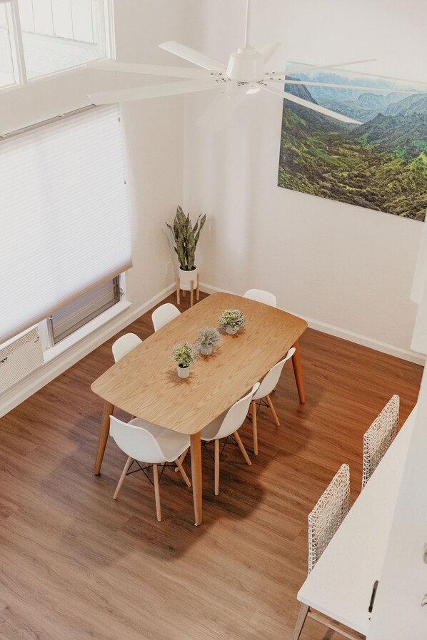 Un comedor moderno con una mesa de madera, sillas blancas, un ventilador de techo, una planta en maceta y un cuadro de un paisaje pintoresco en la pared.