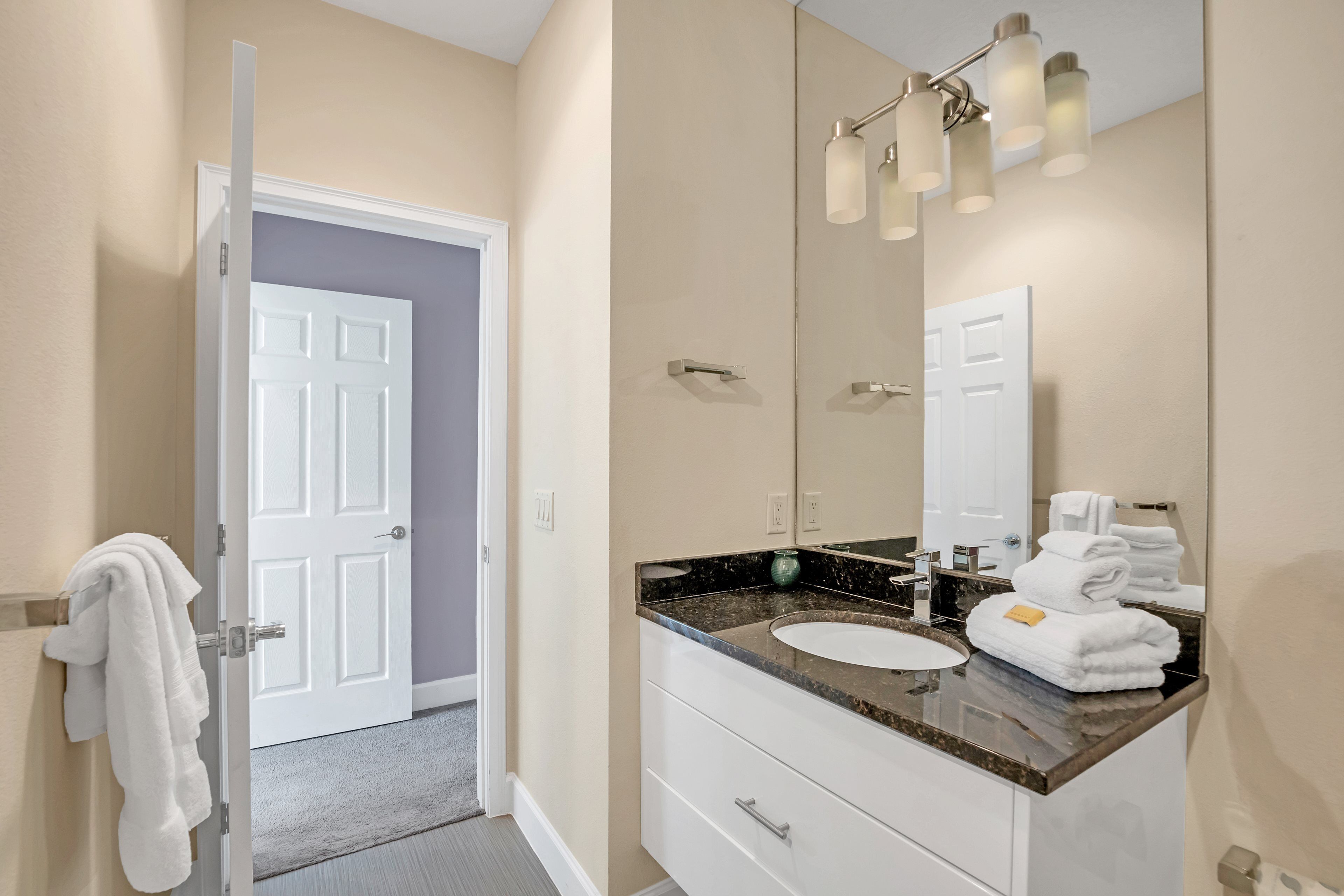 A bathroom with a white door, a sink with a marble countertop, and a mirror with lights above it.