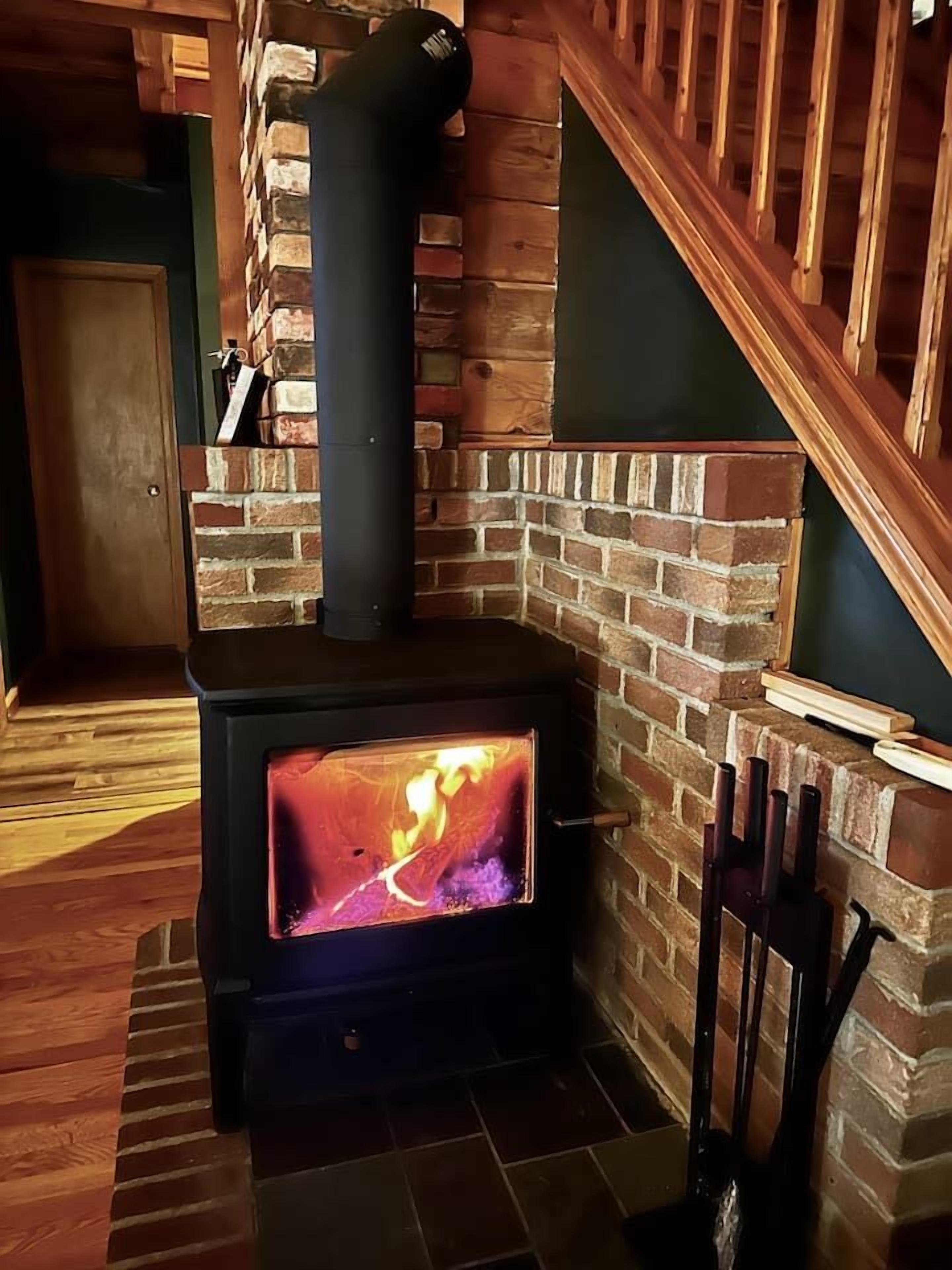 A wood-burning stove with a glass front, set in a corner with a brick wall and wooden staircase.
