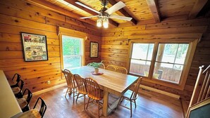Una cabaña de madera con comedor, que incluye una mesa grande y sillas, un ventilador de techo y ventanas amplias.