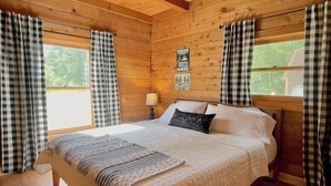 Un dormitorio de cabaña de madera con una cama grande, cortinas a cuadros y una ventana con vista a follaje.