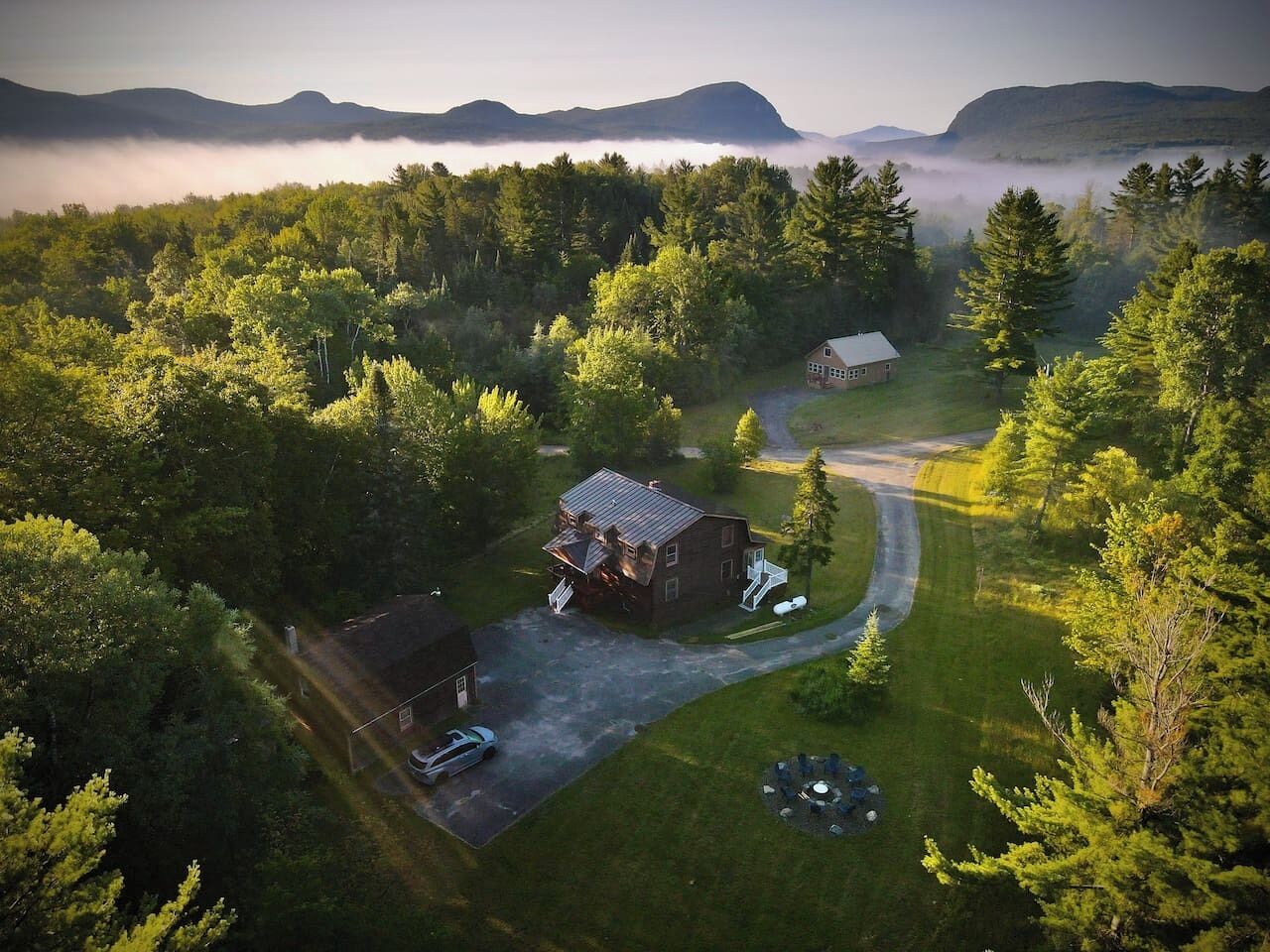 An aerial view of a rural property with two main buildings, a driveway, and a surrounding forest.