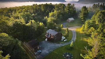 An aerial view of a rural property with two main buildings, a driveway, and a surrounding forest.