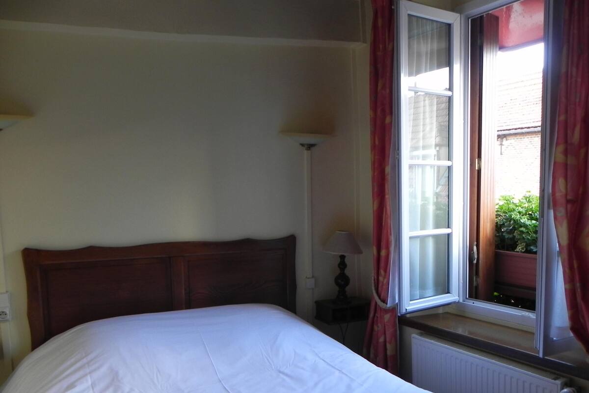 A bedroom with a wooden headboard, a white bedspread, a bedside lamp, and a window with red curtains.