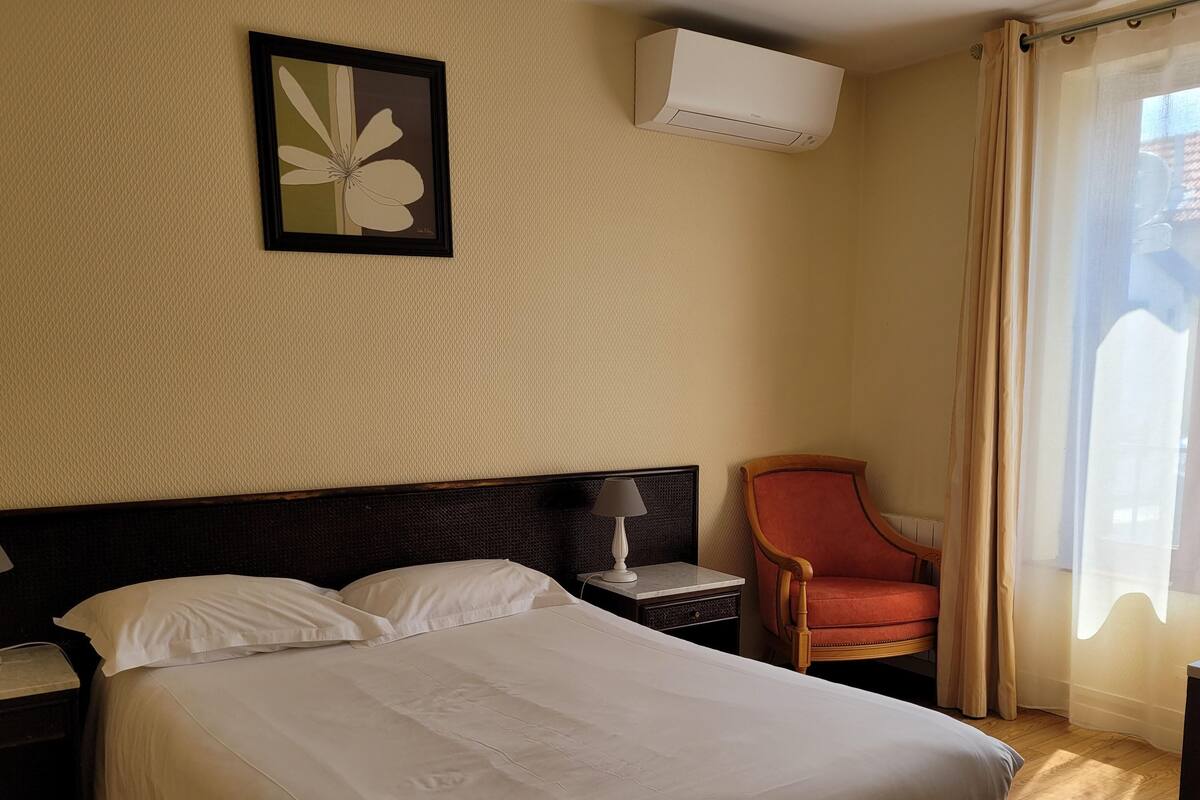 A hotel room with a bed, a nightstand with a lamp, a chair, a framed picture on the wall, and an air conditioning unit.