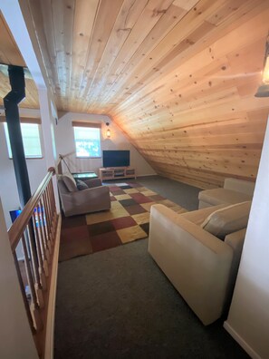 Habitación estilo loft con techo inclinado, un sofá, un televisor y una alfombra a cuadros.