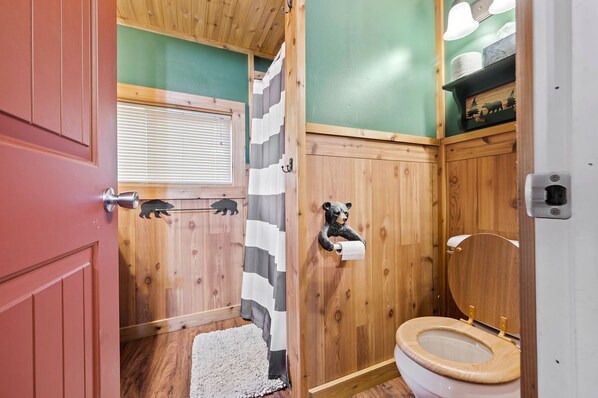 A compact bathroom with wooden walls, a striped shower curtain, a toilet with a wooden seat, and a bear-shaped toilet paper holder.
