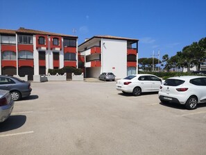 Um parque de estacionamento com vários carros, um edifício de vários andares com fachada vermelha e branca, e um céu azul claro.