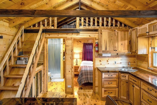 Interior de una cabaña con escalera de madera, cama, pequeña cocina y baño.
