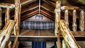 Une chambre à coucher aménagée dans un grenier, avec un plafond en pente, des poutres en bois, une couvre-lit à carreaux et une fenêtre avec des rideaux.