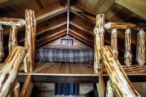 Dormitorio tipo loft con techo inclinado, vigas de madera, colcha a cuadros y ventana con cortinas.