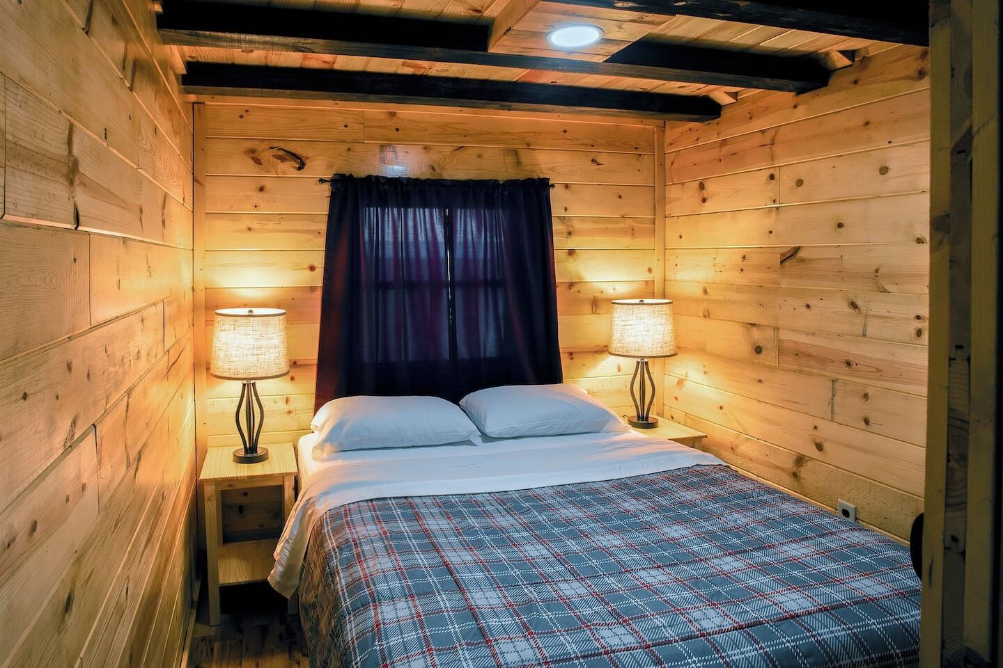 A wooden cabin room with a bed, two bedside lamps, and a dark curtain.