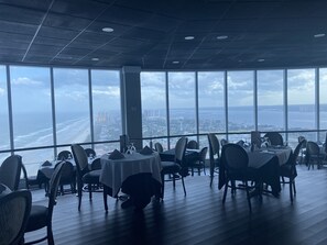 A restaurant with large windows offering a view of the beach and city skyline. Tables are set with chairs and table settings.