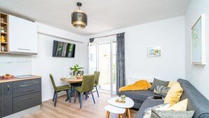 Een moderne woonkamer met een grijze bank, een eettafel met stoelen, een klein wit bijzettafeltje, en een keukenhoek met grijze kasten en een houten aanrecht.