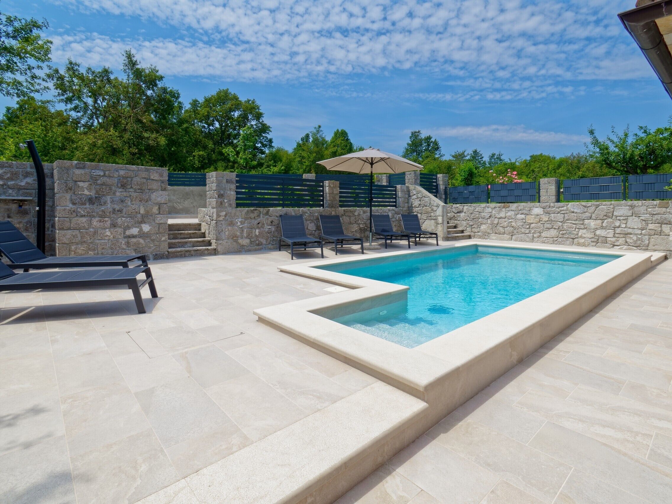 A rectangular swimming pool with a stone border, surrounded by a stone wall and a wooden fence. There are lounge chairs and a shaded area with an umbrella.