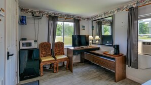 A compact hotel room with a wooden desk, two chairs, a television, a microwave, and a large window with curtains. - The Blue Cottage@ Twin Hills Cottages (Ellsworth)