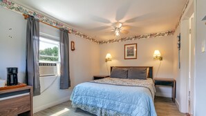 A bedroom with a bed, two bedside lamps, a ceiling fan, a window with curtains, a nightstand, and a desk with a coffee maker. - The Blue Cottage@ Twin Hills Cottages (Ellsworth)