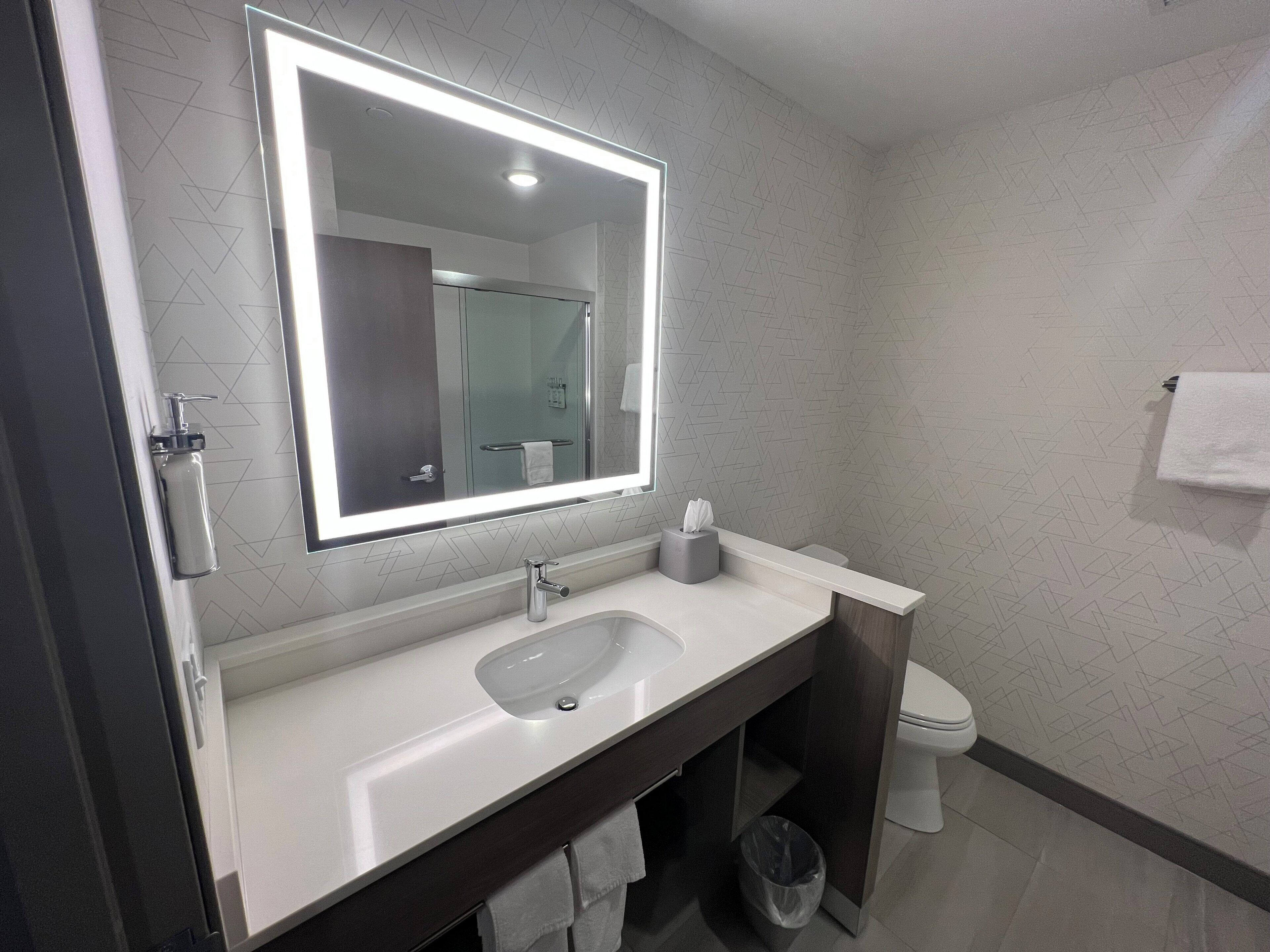 A modern bathroom with a large mirror, a sink, a toilet, and a trash bin.