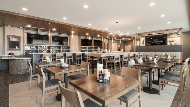 A modern restaurant with wooden tables and chairs, a bar area, and a large TV screen.