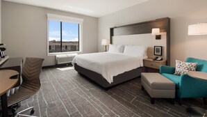 A hotel room with a large bed, a desk, a chair, a sofa, and a window with a view of buildings.