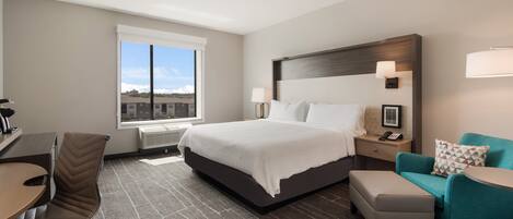 A hotel room with a large bed, a desk, a chair, a sofa, and a window with a view of buildings.