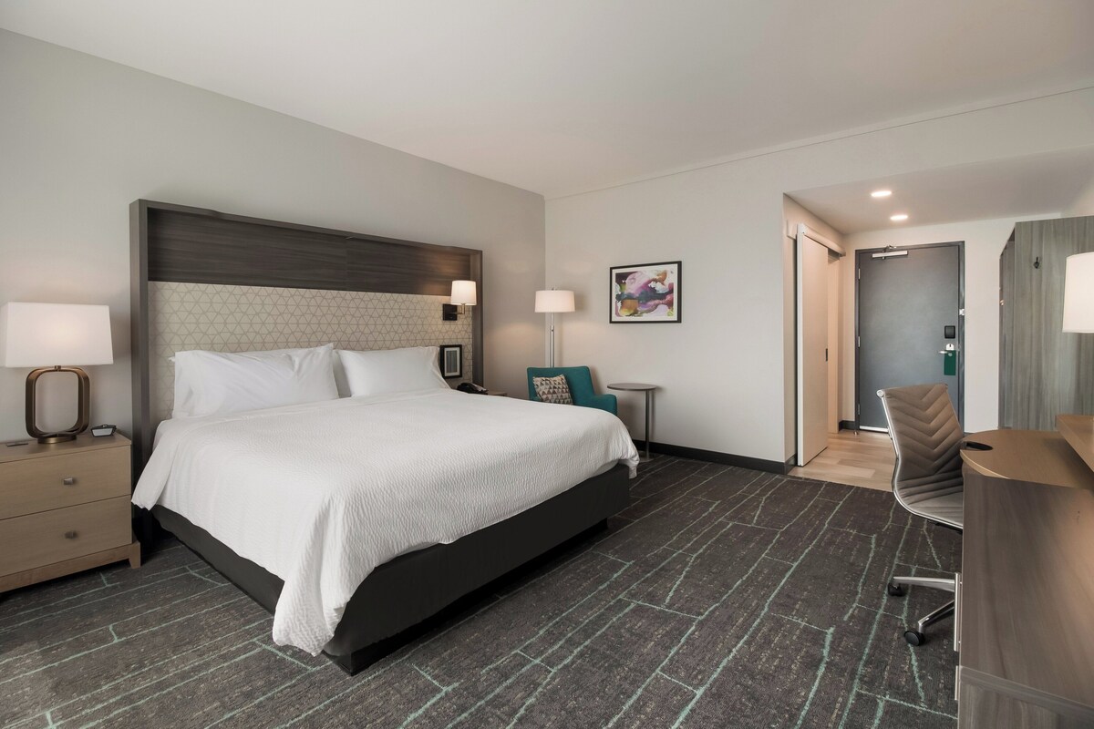 Une chambre d’hôtel avec un grand lit, un bureau, une chaise, une table de chevet, une lampe et un tableau au mur.