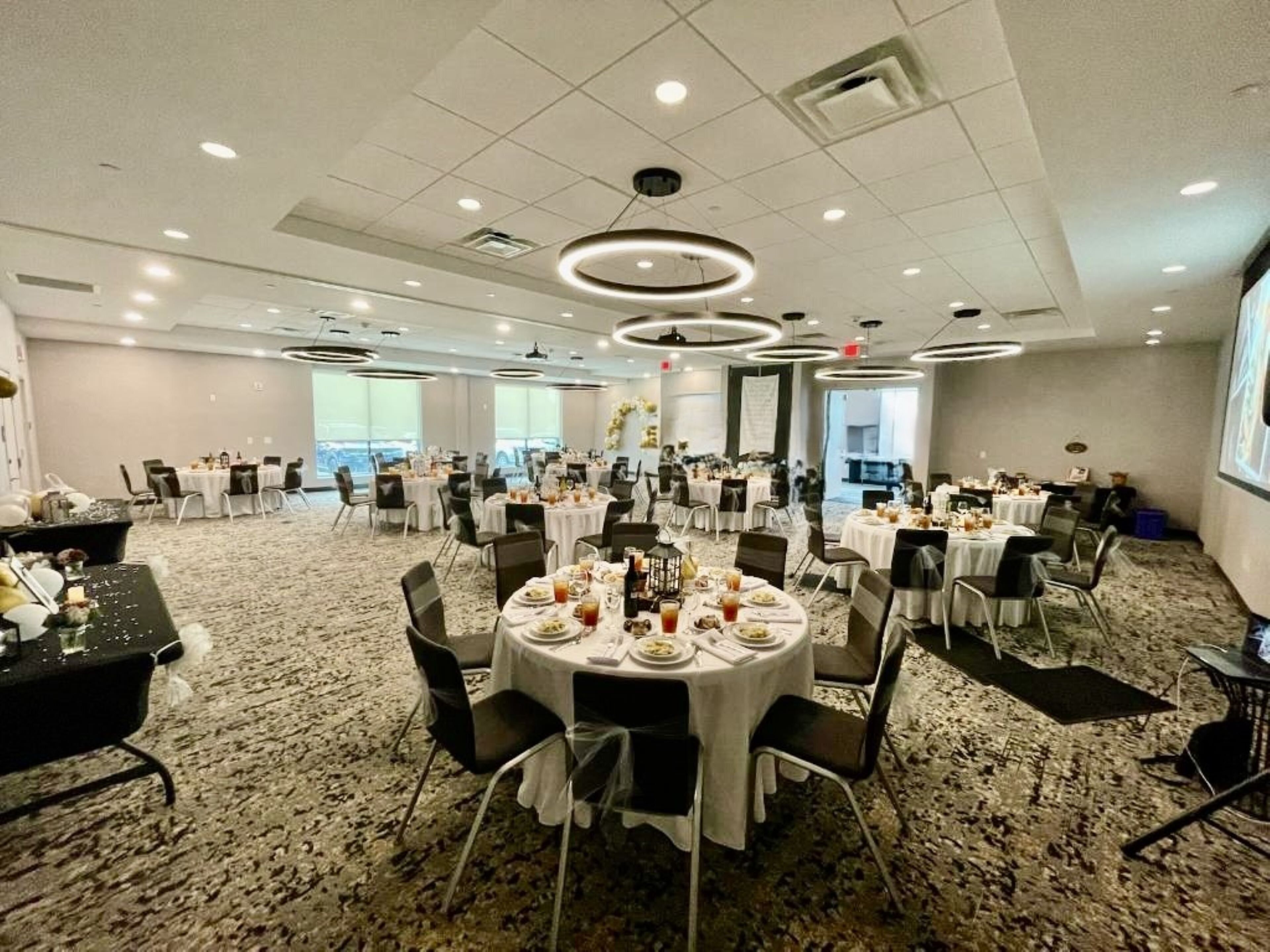 A spacious banquet hall with round tables set for dining, carpeted flooring, and a high ceiling with recessed lighting.