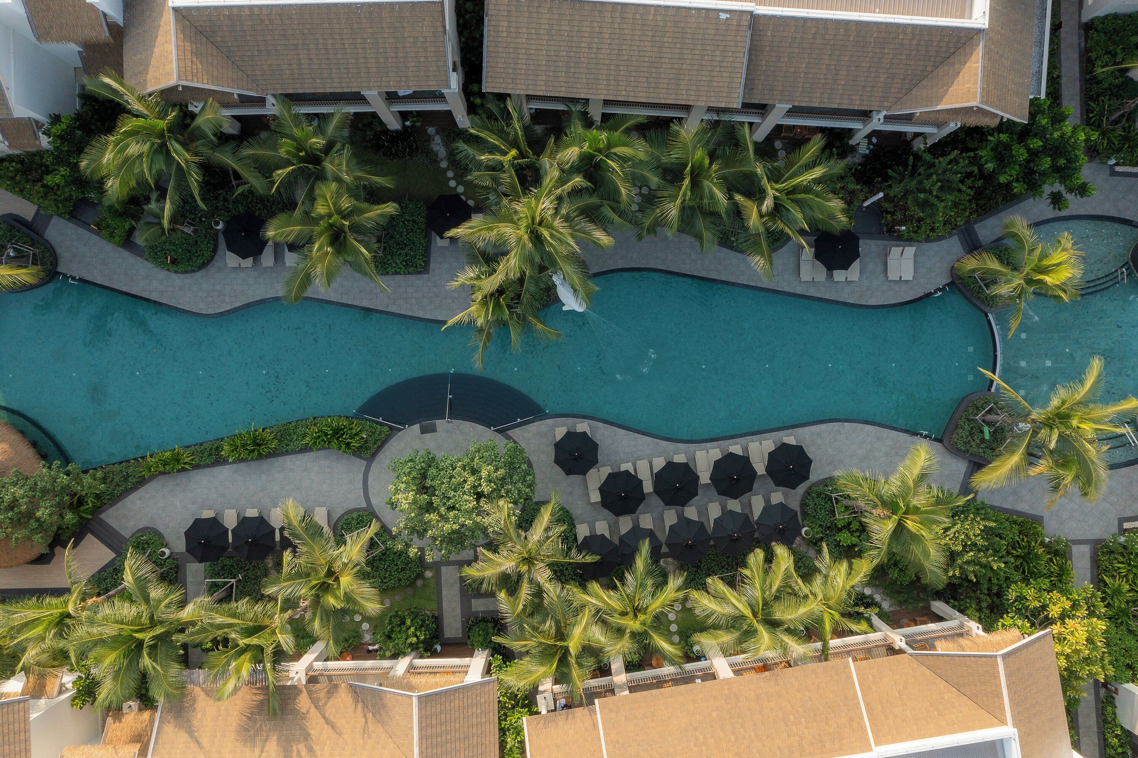 An aerial view of a resort with a swimming pool, palm trees, and outdoor seating areas.